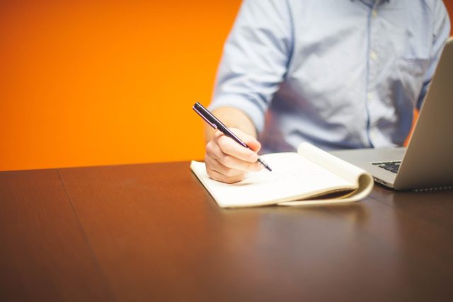 Osoba pisząca notatki w notesie przy laptopie podczas planowania biznesu.